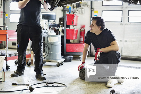 Junger Mechaniker,  der in der Werkstatt auf dem Reifen sitzt und einen Mitarbeiter beim Reparieren des Autos ansieht
