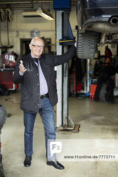 Portrait of happy senior man gesturing while standing by car jack in auto repair shop