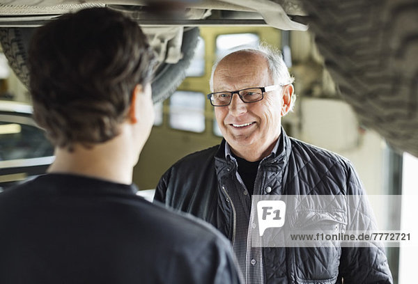 Glücklicher älterer Mann  der den Mechaniker unter dem Auto in der Autowerkstatt ansieht.