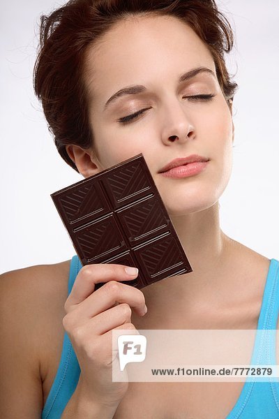 Porträt einer jungen brünetten Frau mit geschlossenen Augen  die eine Tafel Schokolade gegen das Gesicht hält.