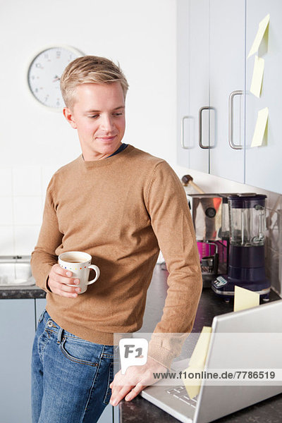 Junger Mann in der Küche mit Kaffeetasse und Laptop