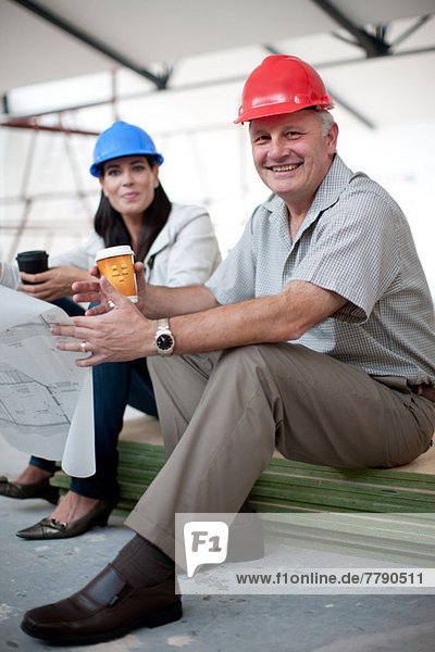 Lächelnde Bauarbeiterinnen und Bauarbeiter mit Kaffee und Blaupause im Sitzen