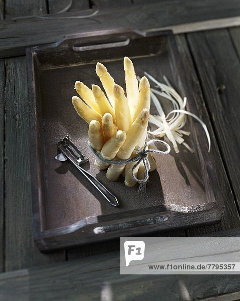 A bunch of white asparagus with a peeler on a wooden tray