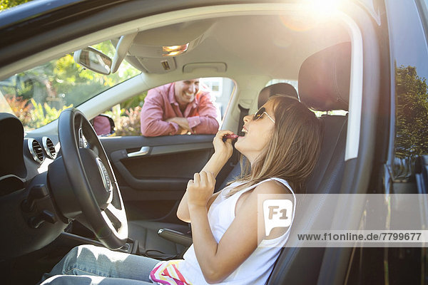 Young girl applying Lip Gloss  pretending to be old enough to drive as her smiling father watches on on a sunny summer evening in Portland  Oregon  USA