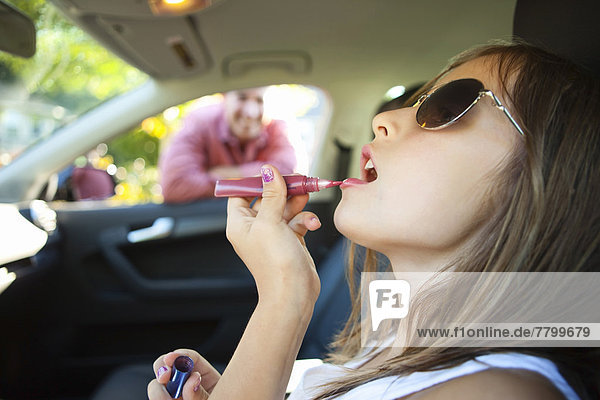 Young girl applying lip glss pretending to be old enough to drive as her smiling father watches on on a sunny summer evening in Portland  Oregon  USA