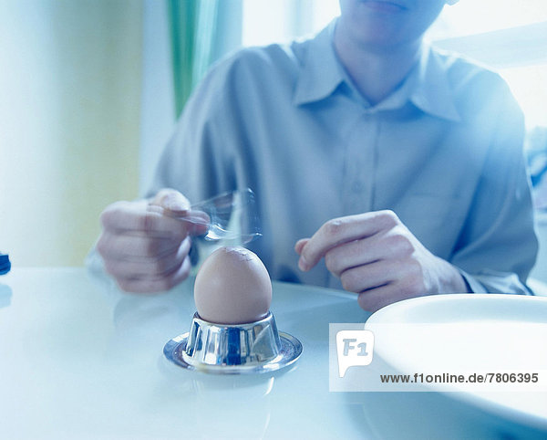 Mann sitzt am Tisch und klopft Frühstücksei mit Löffel auf