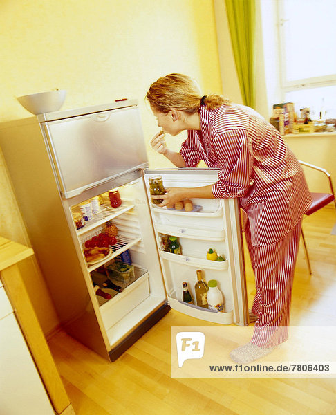 Frau im Schlafanzug steht am geöffneten Kühlschrank und isst Essiggurken