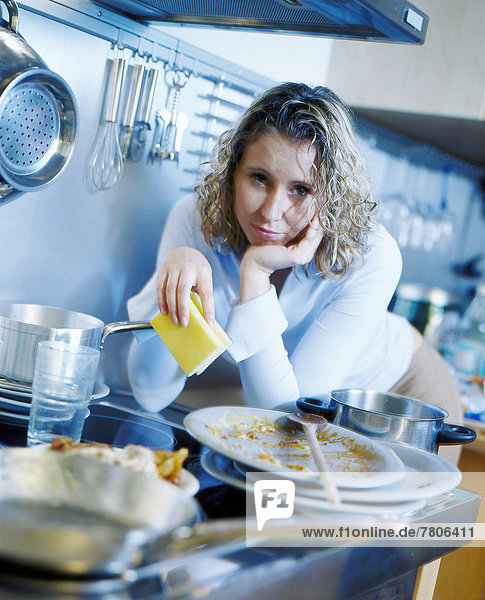 Genervte Frau mit Spülschwamm steht in der Küche vor schmutzigem Geschirr