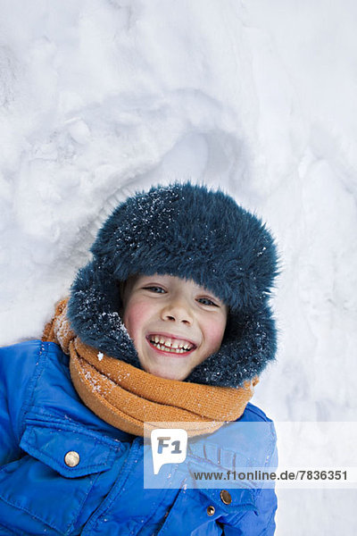 A young cheerful boy wearing warm clothing outdoors lying in the snow