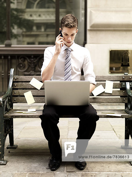 Geschäftsmann  der ein Smartphone benutzt  während er einen Laptop benutzt und auf einer Bank mit Haftnotizen sitzt.