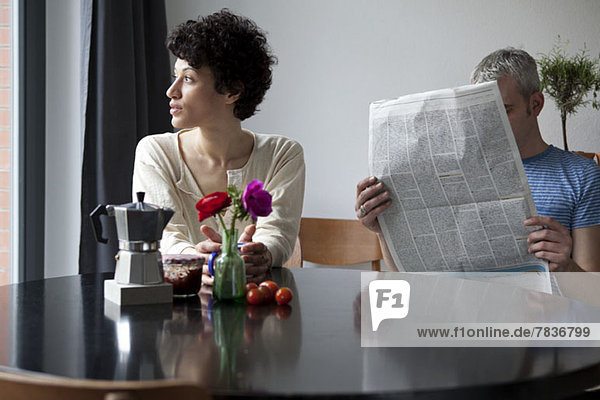 Eine ernste Frau schaut aus dem Fenster  während sich ihr Freund hinter einer Zeitung versteckt.