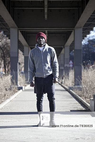 Ein hipper junger Mann steht in Sportausrüstung auf einem Fußweg unter einer Brücke