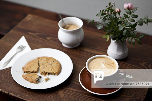 Ein Keks  Café Latte  ein Glas Zucker und eine Vase mit Blumen auf einem Tisch in einem Café.