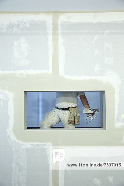 Ein Hausmaler hält eine Kelle  gesehen durch ein Fensterloch in einer neu errichteten Wand.