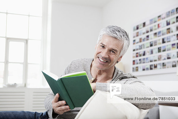 Porträt eines reifen Mannes  der das Buch auf der Couch liest  lächelnd