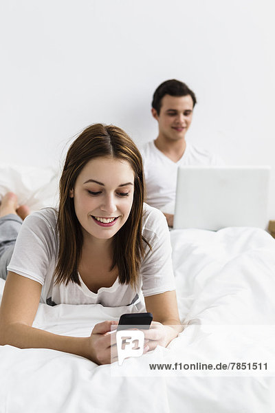 Junge Frau mit Handy  junger Mann mit Laptop im Hintergrund  lächelnd