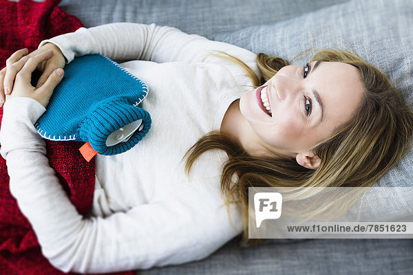 Porträt einer jungen Frau auf der Couch liegend mit Wärmflasche  lächelnd