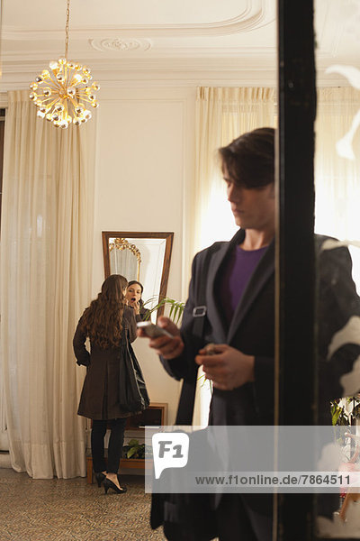 Man waiting by door as woman in background applies lipstick in mirror