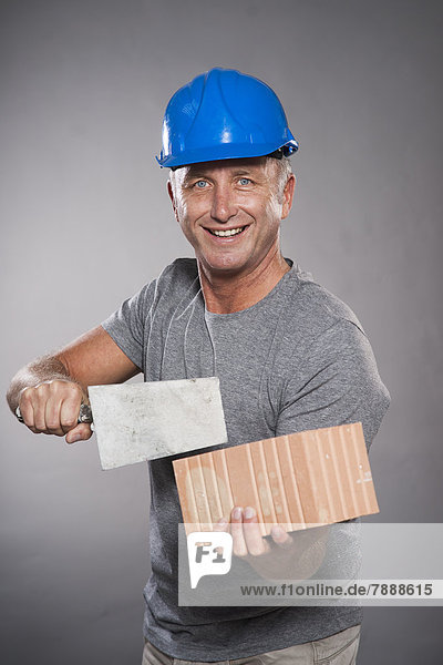 Lächelnder reifer Mann mit Bauarbeiterhelm  Maurerkelle und Ziegelstein