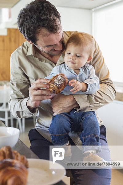 Man feeding bread to his son at a kitchen counter