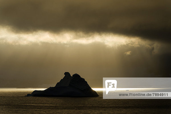 Sonnenlicht scheint durch Wolkenlücke  davor Silhouette eines Eisberges