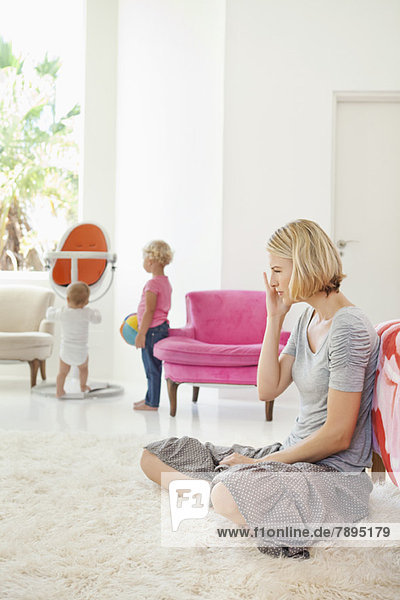 Frau sitzt auf einem Teppich und ihre Kinder spielen im Hintergrund