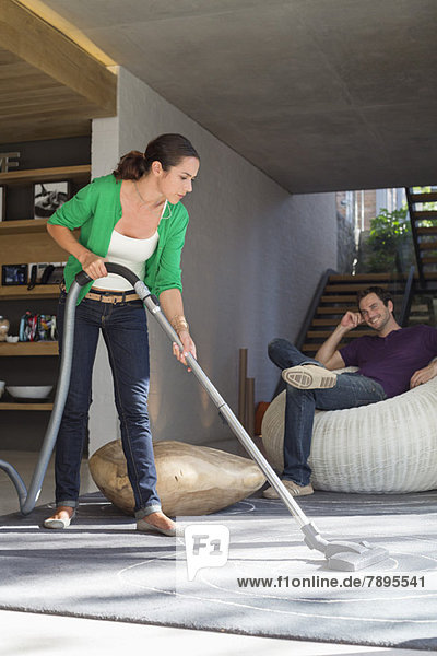 Woman cleaning house with a vacuum cleaner with her husband sitting on a seat