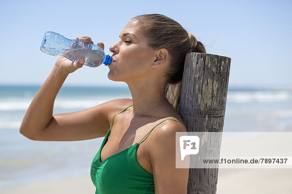 Woman leaning against a wooden post on the beach drinking water