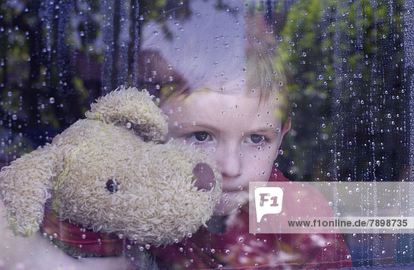 Junge schaut aus Fensterscheibe mit Wassertropfen  Kuscheltier im Arm