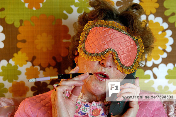 Alte Frau mit einer Augenmaske zum Schlafen und Zigarettenhalter im bunten Retro-Look