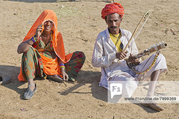 Inder mit Turban und dem traditionellen Beinkleid Dhoti  spielt auf einer Sitar  eine Frau mit buntem Sari hockt daneben