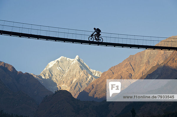 Cyclist crossing a suspension Bridge over the Kali Ghandaki Valley  Nilgiri Mountain at back