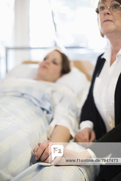 Ältere Frau schaut weg  während sie die Hand der Tochter auf dem Bett in der Krankenstation hält.