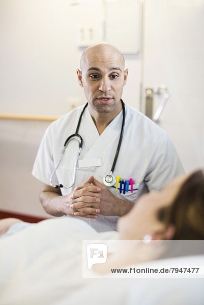 Mittelgroßer männlicher Arzt  der mit einer Patientin kommuniziert  die auf dem Bett in der Krankenstation liegt.