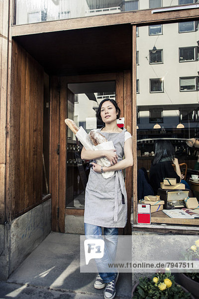 Portrait of confident young female owner holding bread loafs while standing outside bakery