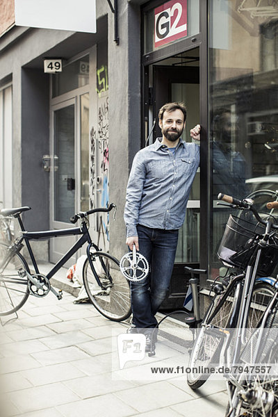 Porträt des mittleren erwachsenen männlichen Besitzers mit Pedal am Eingang der Fahrradwerkstatt
