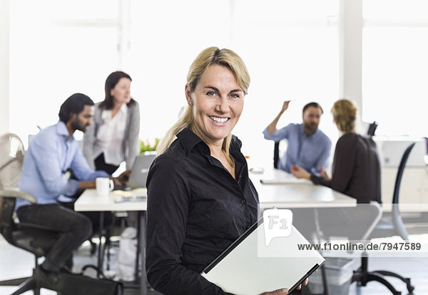 Porträt einer reifen Geschäftsfrau mit Laptop und Kollegen im Hintergrund im Büro