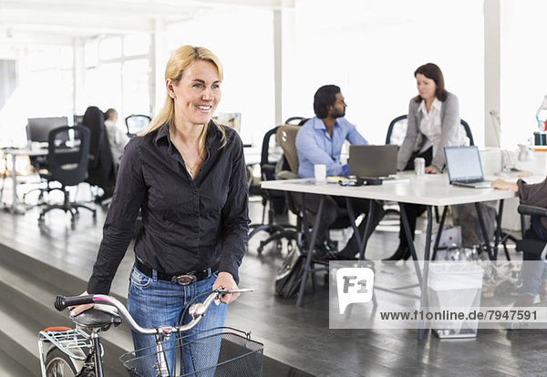 Mature businesswoman looking away while holding bicycle with colleagues working in background at office