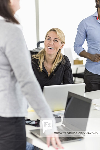Happy mid adult businesswoman looking at colleague at desk in office
