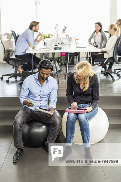 Business people discussing work while sitting on fitness balls with colleagues in background at office