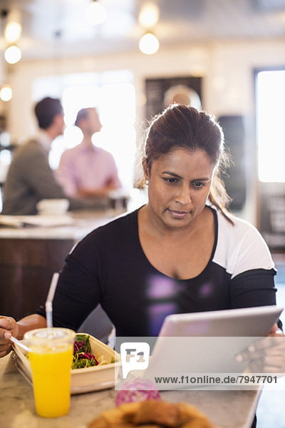 Geschäftsfrau mit digitalem Tablett beim Frühstück am Restauranttisch mit Kollegen im Hintergrund