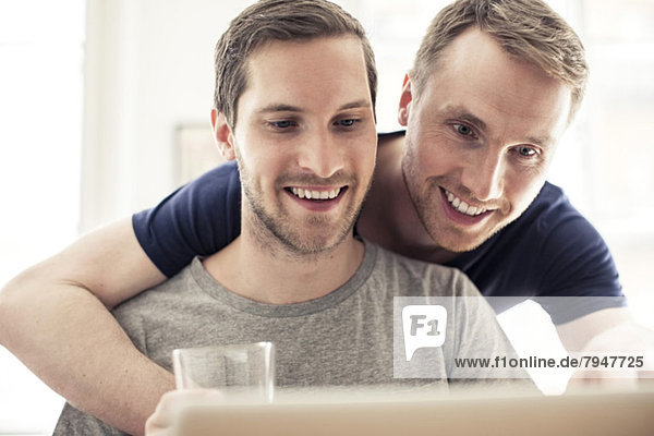 Glückliches homosexuelles Paar  das den Laptop benutzt  während es sich zu Hause umarmt.