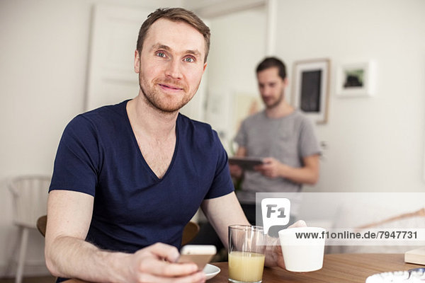 Porträt eines jungen schwulen Mannes mit Handy beim Frühstück am Tisch mit Partner im Hintergrund