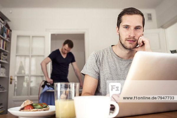 Portrait of gay man sitting with laptop at breakfast table with partner ironing in background