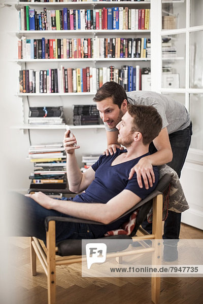 Gay men partner using mobile phone together against bookshelf at home