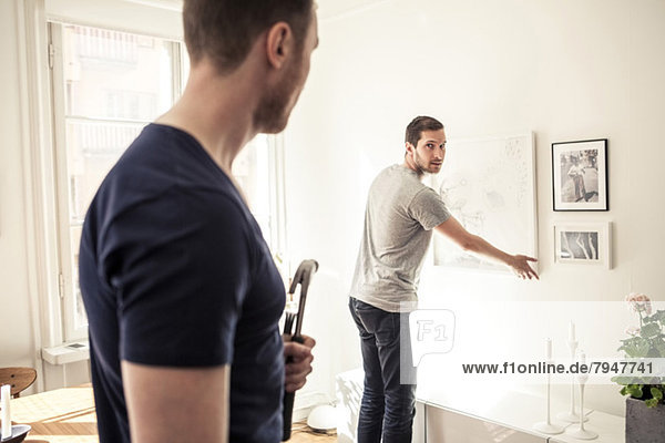 Young gay man looking over shoulder at partner while hanging frame on wall in home