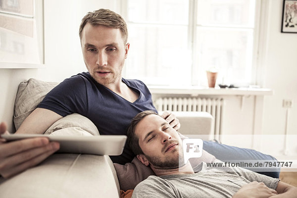 Junger schwuler Mann mit digitalem Tablett  während der Partner zu Hause auf dem Sofa schläft.