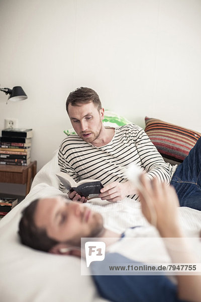Junger schwuler Mann liest Buch  während der Partner zu Hause im Vordergrund Musik hört.