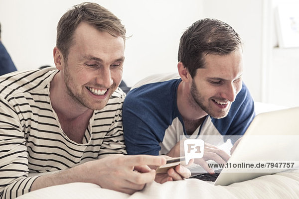 Happy gay men using credit card and laptop to shop online while lying in bed at home