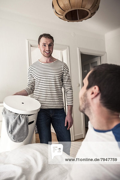 Junger schwuler Mann  der den Partner ansieht  während er den Wäschekorb zu Hause hält.